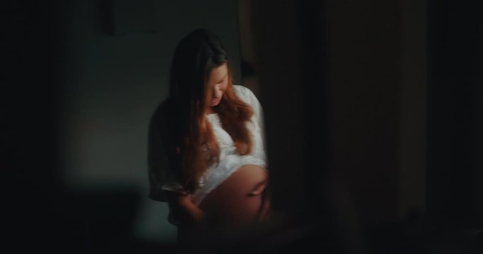 Authentic shot of an young pregnant woman in white dress is caressing her belly just woke up in the morning in a bedroom.