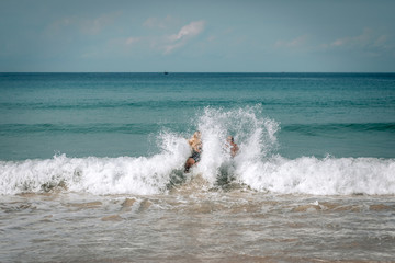 Handsome young couple standing on a beach in Phuket Island, Thailand, getting hit by big waves splash - Image