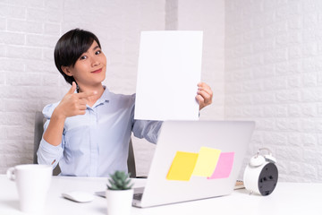 Happy woman sitting at home office and showing blank sheet