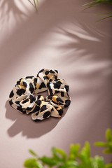 Object photo of a with beige scrunchie with leopard print. The scrunchie is lying on a beige background. There are leaves in corners of a photo. The scrunchie and leaves casting a shadow.