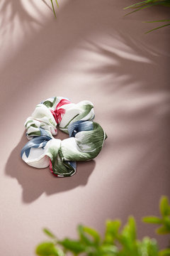 Object photo of a white scrunchie with flower ornament. The scrunchie is lying on a beige background. There are leaves in corners of a photo. The scrunchie and leaves casting a shadow.