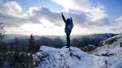 Man on top of the cliff raised his hands joyfully. The view from the back. Sunlight through the clouds illuminates the mountains. Winter snow.