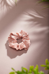 Object photo of a pale pink scrunchie with white polka dot. The scrunchie is lying on a beige background. There are leaves in corners of a photo. The scrunchie and leaves casting a shadow.