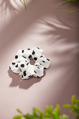 Object photo of a white scrunchie with black polka dot. The scrunchie is lying on a beige background. There are leaves in corners of a photo. The scrunchie and leaves casting a shadow.