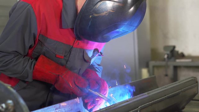 Man in gloves and welding helmet works with metal, high temperature welding, argon welding, sparks, blue glow, hard work, factory production, employee of welding workshop at work, complex product