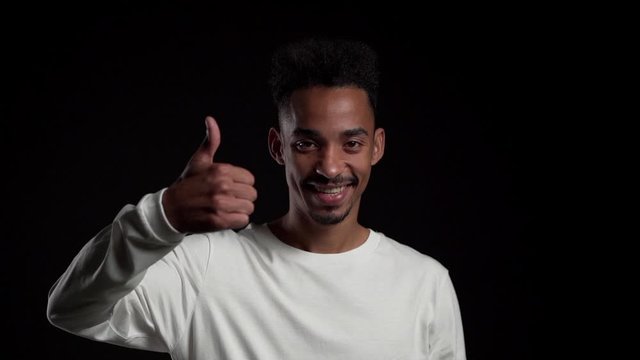 Handsome man in white wear on black studio background smiles to camera and gives thumbs up. Happy african american guy showing gesture of approval. Winner.Success.