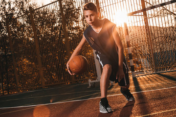 Sports and basketball. A young teenager in a black tracksuit is playing a basketball. Sunset light
