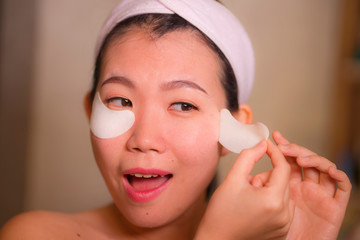 young beautiful and happy Asian Japanese woman smiling to bathroom mirror applying hydrating eye patches under the eyes in beauty cosmetics and facial skin care