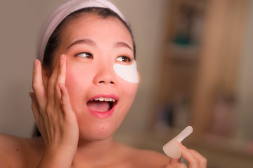 young beautiful and happy Asian Japanese woman smiling to bathroom mirror applying hydrating eye...