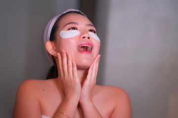 young beautiful and happy Asian Korean woman smiling to bathroom mirror applying hydrating eye...