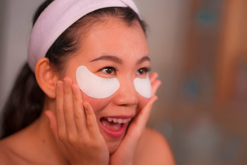 young beautiful and happy Asian Korean woman smiling to bathroom mirror applying hydrating eye...