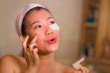 young beautiful and happy Asian Korean woman smiling to bathroom mirror applying hydrating eye...