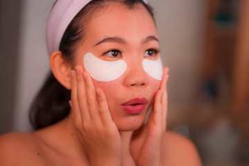 young beautiful and happy Asian Chinese woman smiling to bathroom mirror applying hydrating eye...