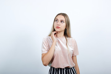 Portrait of young woman in pastel pink shirt hearing gesture, looking aside, keeping hand on chin over isolated white wall background. People sincere emotions, lifestyle concept
