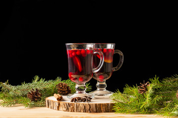 Two glasses with homemade mulled wine on a wooden saw. Black background. Real spruce branches. Fir cones.