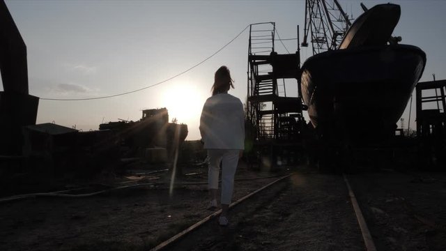lady silhouette walks along old seaport crossing railway with old ship against golden sundown slow motion backside view