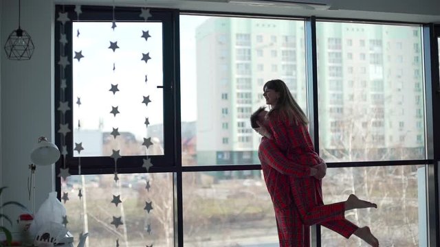 Happy Couple in Red Pajamas Enjoying Time Together at Home. They Hugging and Kissing in Front of Window. City Steet is on the Background. Lifestyle, Romantic Relationships and People Concept