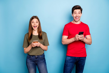 Portrait of amazed crazy two married people use smart phone read social network newas impressed got like incredible feedback wear green t-shirt denim jeans isolated blue color background - obrazy, fototapety, plakaty