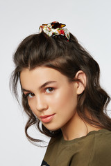 Naklejka premium Closeup half-turn face portrait of girl with dark wavy hair, wearing khaki t-shirt. Her hair is pulling with brown hairpin crab with white bow with red flower ornament. 