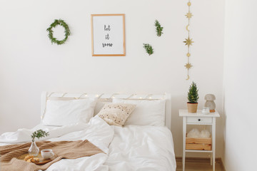 New year winter home interior decor. Holiday decorated room. Stylish cozy white scandinavian bedroom: bed, little christmas tree in a pot on bedside table, wreath, pine branches, led garland lights.