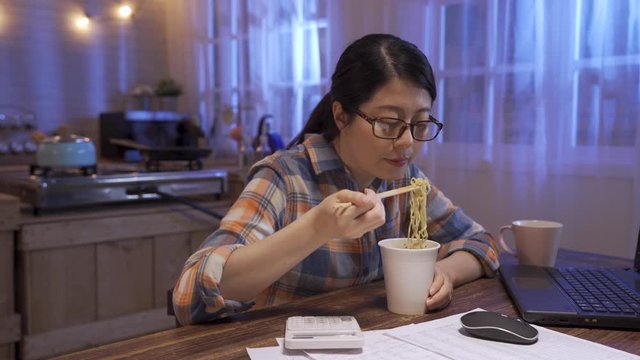 beautiful woman eating yummy hot and spicy instant noodle in cup using chopsticks in night home kitchen. Asian girl servile end of month with cheap fast food. female enjoy unhealthy late night supper