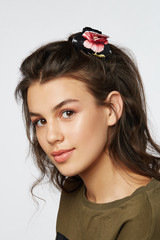 Naklejka premium Closeup half-turn face portrait of girl with dark wavy hair, wearing khaki t-shirt. Her hair is pulling with black hairpin crab with red poppy flower painting. Girl is looking straight and smiling. 