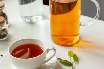 Cup of hot black tea on white table with dry tea jar