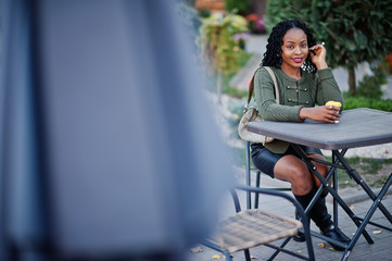 Stylish fashionable african american women in green sweater and black skirt posed outdoor cafe, sitting by table with cup of coffee.