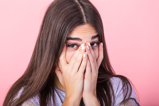 Photo of frightened woman with brown hair in basic t-shirt keeping hands over her face and looking at camera isolated over pink background