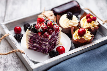 a piece of pancake cake with a cap of berries doused with white chocolate on a plate with Belgian muffins and a bottle of syrup on a tray on a white wooden table