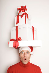 Young man in Santa hat and red turtleneck balancing a tower of Christmas presents on his head