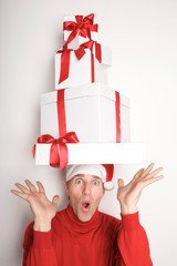 Man in Santa hat and red turtleneck balancing a tower of Christmas presents on his head