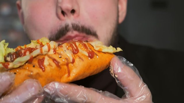Extra large young man eating a hot dog in transparent gloves in a cafe