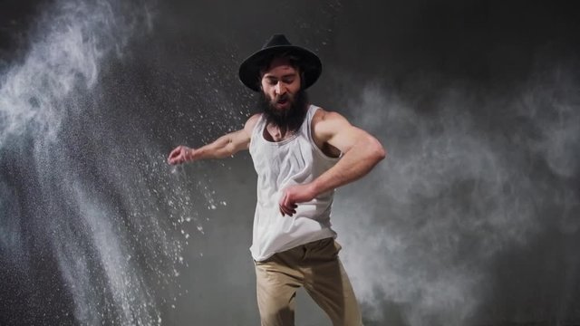 Beard guy a bboying style dancer does difficult tricks on the floor in a studio filled with flour on a black background.