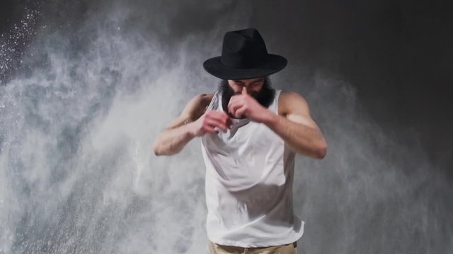 Beard guy a bboying style dancer does difficult tricks on the floor in a studio filled with flour on a black background.
