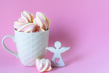 White cup of chocolate with pink marshmallows and a white angel figurine on a pink background. Copy space.