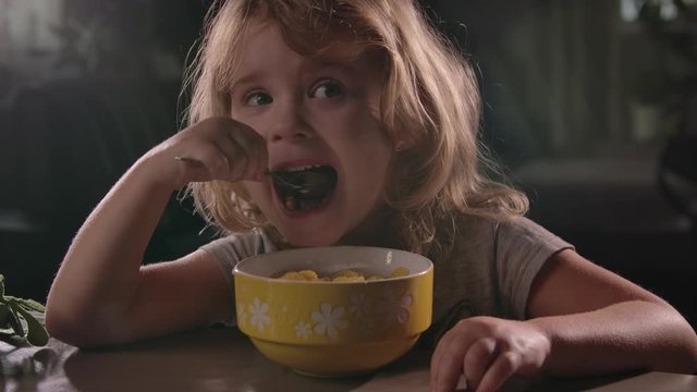 little beautiful hungry girl sits at a wooden table and eats a large spoon from a yellow Cup of cornflakes with milk. A healthy Breakfast is served every morning. a portion of vitamins for the child;