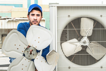 a professional electrician man is showing up the broken fans of heavy unit of an air conditioner at the roof top of a building and wearing blue uniform and head cap