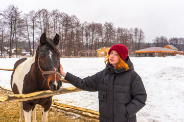 Teen redhead girl patting horse at ranch in snowy day. Winter weekend at farm, trip to countryside....