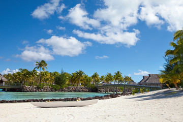 white traditional Polynesian boat on a sandy beach, a bridge across the sea strait to the shore with palm trees and authentic Polynesian thatched roof houses. Polynesia, Tahiti