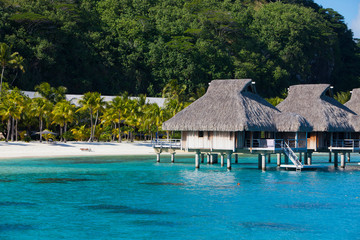 A view from the blue tropical sea of ​​authentic traditional Polynesian houses with a thatched roof over the water and a sandy beach with palm trees and a mountain in the background. Polynesia, Tahiti