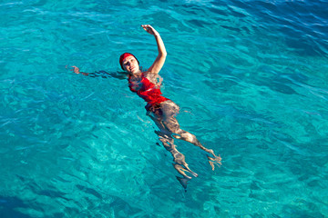 young woman in a red swimsuit and a red sports cap with a good slim figure swims on her side in a bright blue tropical sea and looks at the camera