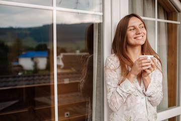 Portrait of a happy smiling young woman in stylish nightwear with a cup of coffee or tea meeting the day standing on the hotel terrace with a gorgeous view of the mountains. place for inscription