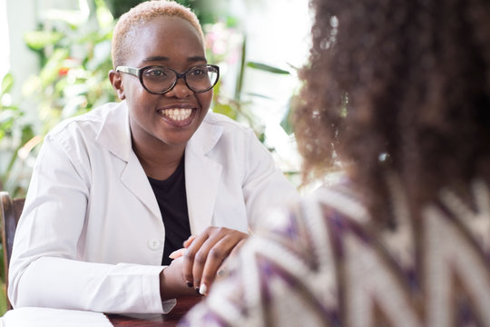 A Young African American Female Doctor With Glasses Shakes Hands With A Patient As A Sign Of Trust Of A Doctor. People Of Mixed Race In The Doctors Office