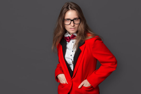girl in white shirt, black vest and red jacket with Burgundy bow tie and classic black pants office style and glasses on gray background looks contemptuously