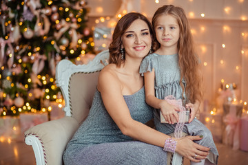 waiting for the New Year holiday! beautiful mother and daughter in elegant silver dresses give each other boxes of gifts. behind them is a Christmas tree with Christmas lights