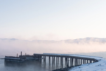 old wooden pier for large ships on the river bank covered in thick white fog from the water at dawn on a frosty winter day outside the Arctic Circle