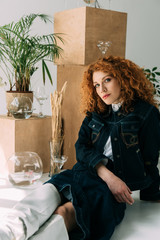 trendy redhead girl sitting and posing near wooden boxes, glasses and plants on grey