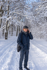 Fototapeta na wymiar Handsome middle-aged man walking in winter snowy park or forest. Attractive man in jacket, scarf and cap talking on mobile phone. Winter mood, authentic lifestyle concept, stylish male outfit