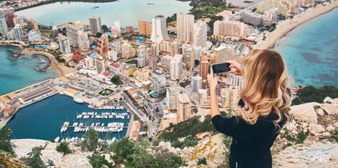 Fototapeta na wymiar Woman climbed up on Penon de Ifach rock. Enjoy view of city from top, stand on peak of mountain takes photo of cityscape and scenery. Tourism, landmark, tourist capture moment concept, Calpe, Spain